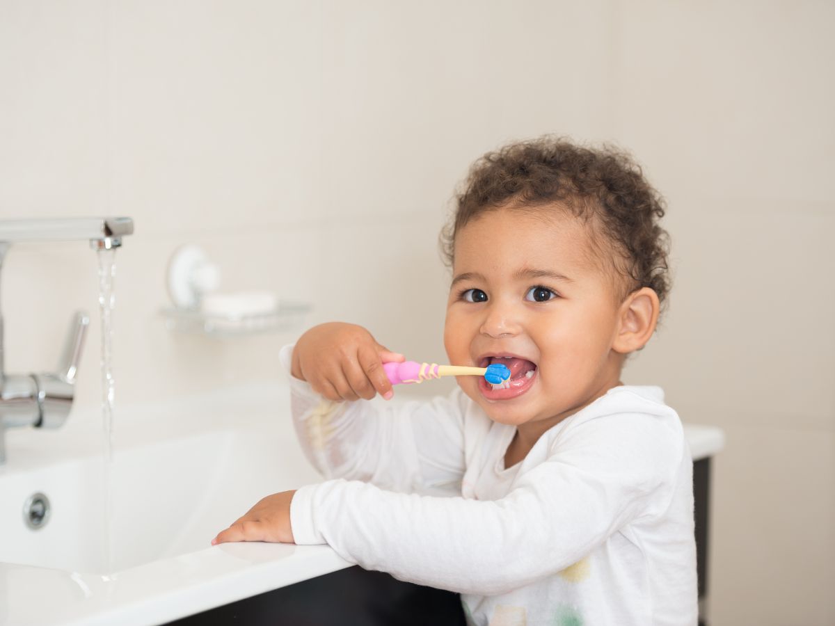 Young child brushing teeth with fluoride toothpaste at bathroom sink, wearing white shirt and smiling.