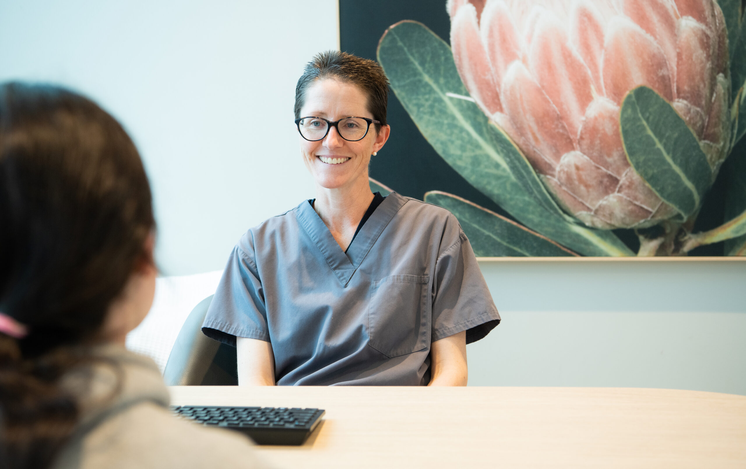  Dr. Sara in scrubs smiles while discussing how to stop grinding teeth, seated across from another person at her desk.