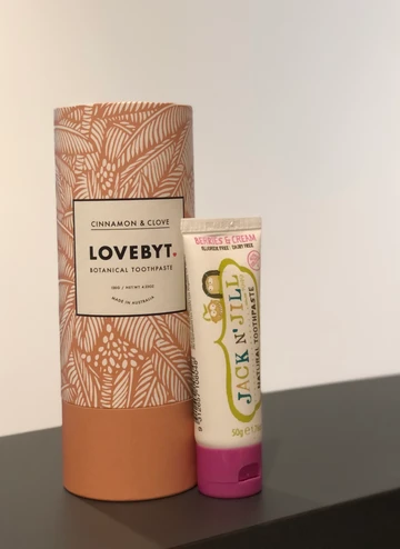 Two toothpaste containers on a dark surface: one large LOVE BYT and one small Jack N’ Jill vegan tube.