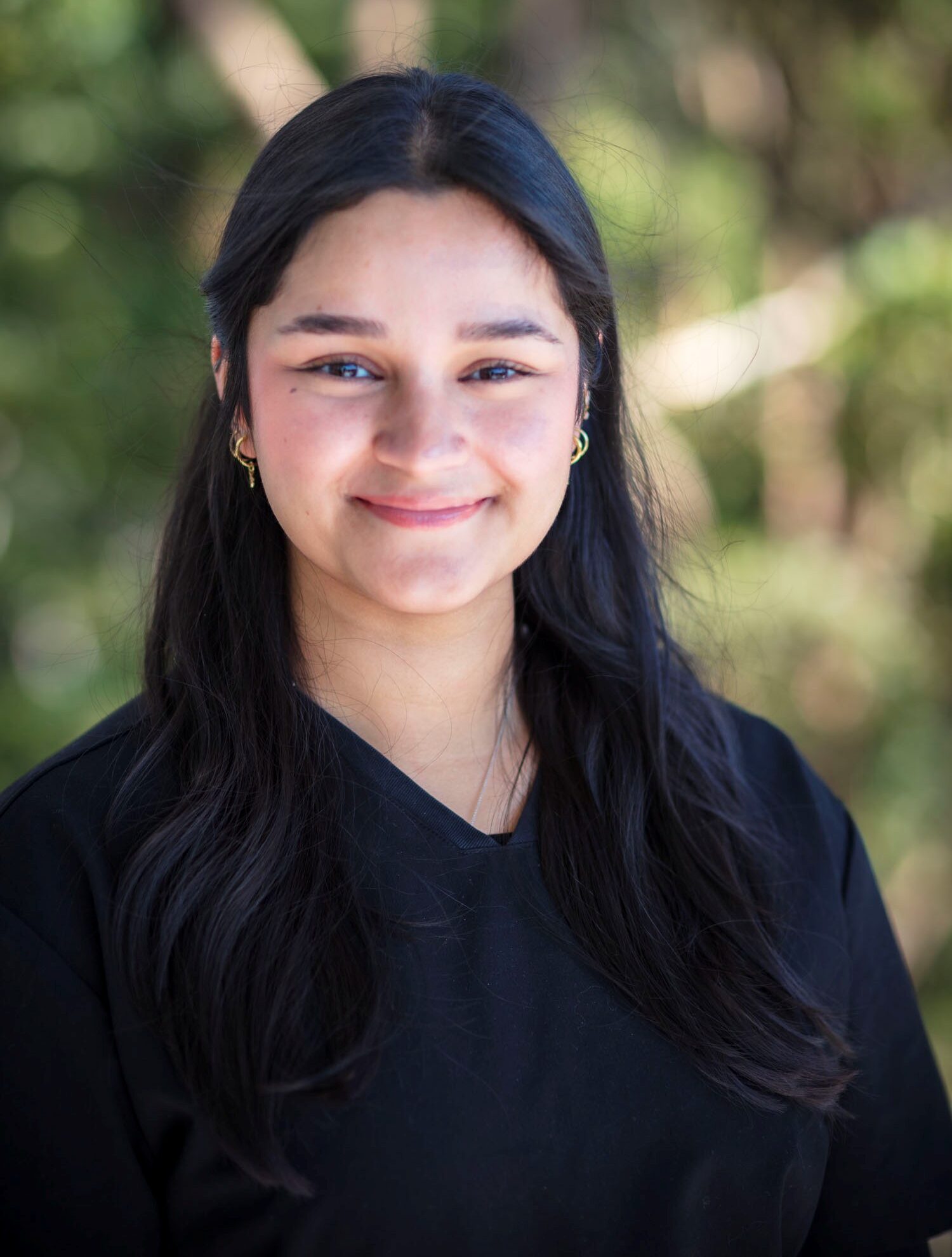 Oditi Sarkar, Tooth Sparkler team member, smiles outdoors in a black top with green blurred background.