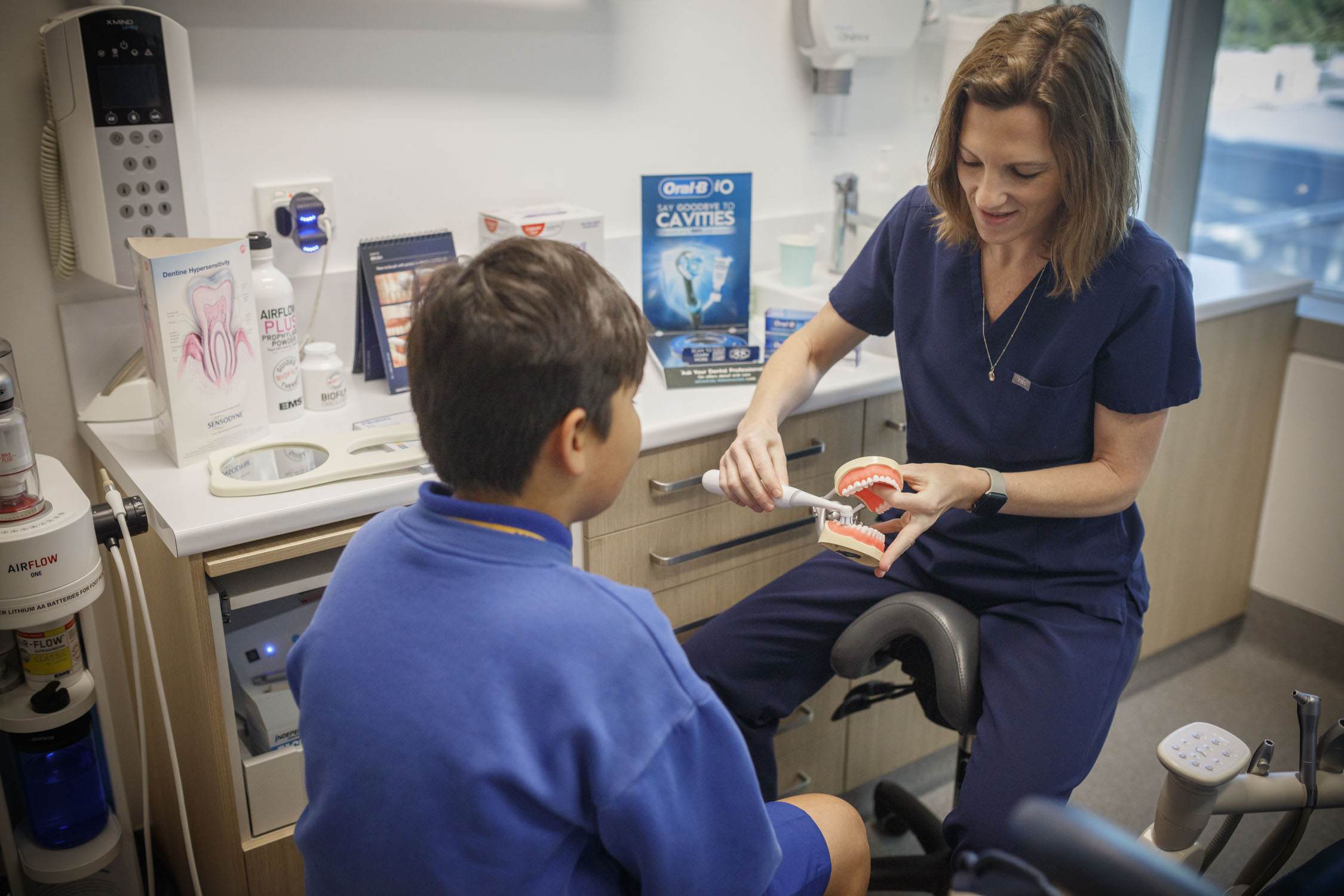 Dentist in scrubs teaches boy to brush teeth with Tooth Sparkler and model jaw; dental posters in background.